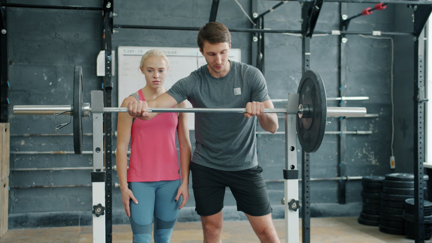 Trainer begeleidt vrouw die barbell-oefening doet in de sportschool.