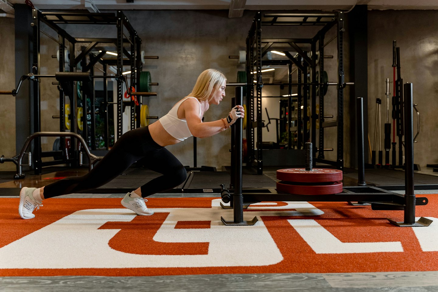 Vrouw in sportkleding duwt een slee met gewichten over de vloer van de sportschool.